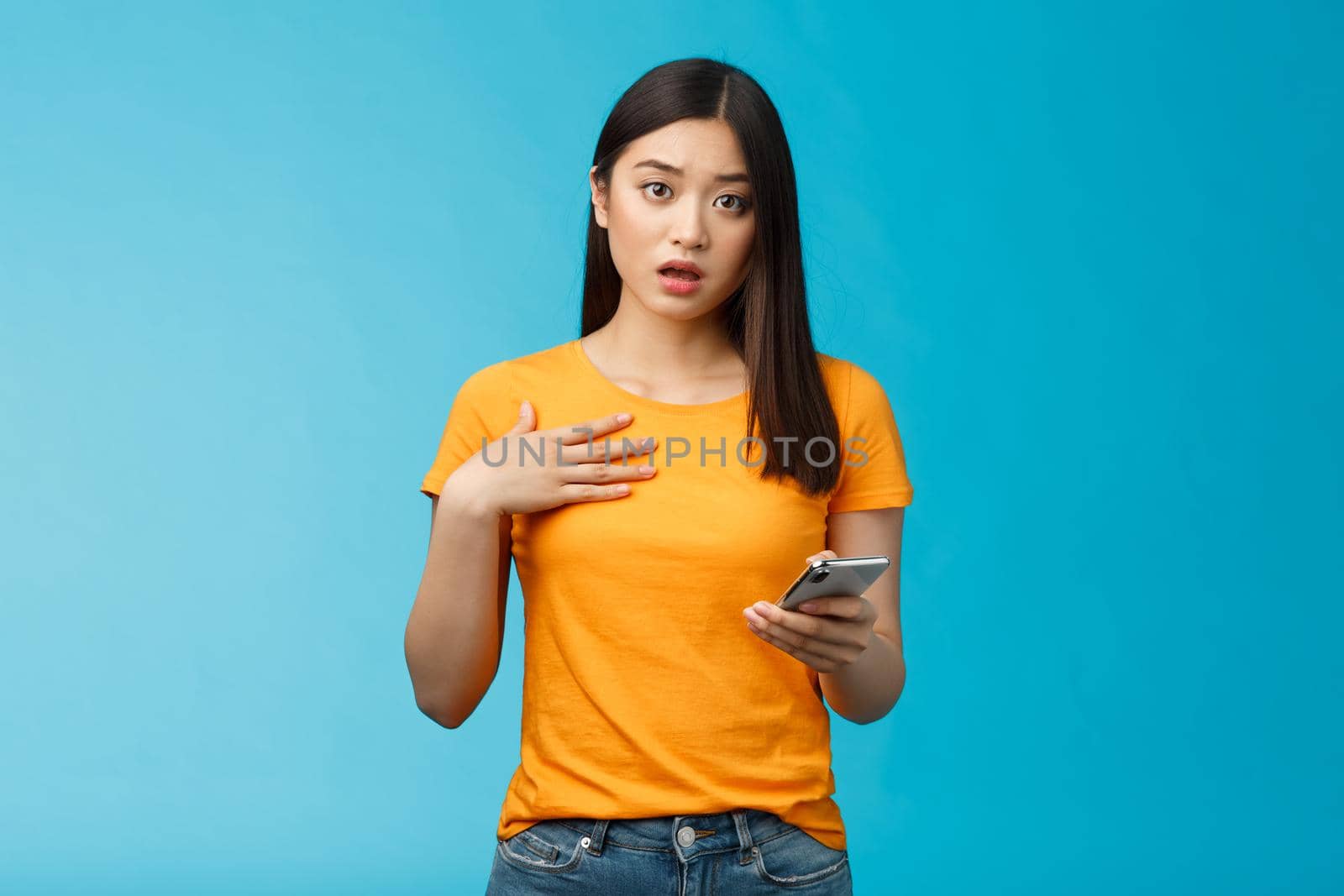 Offended shocked asian girl receive insulting message via internet social media, hold smartphone look surprised upset gasping, drop jaw touch chest shook, stand blue background. Copy space