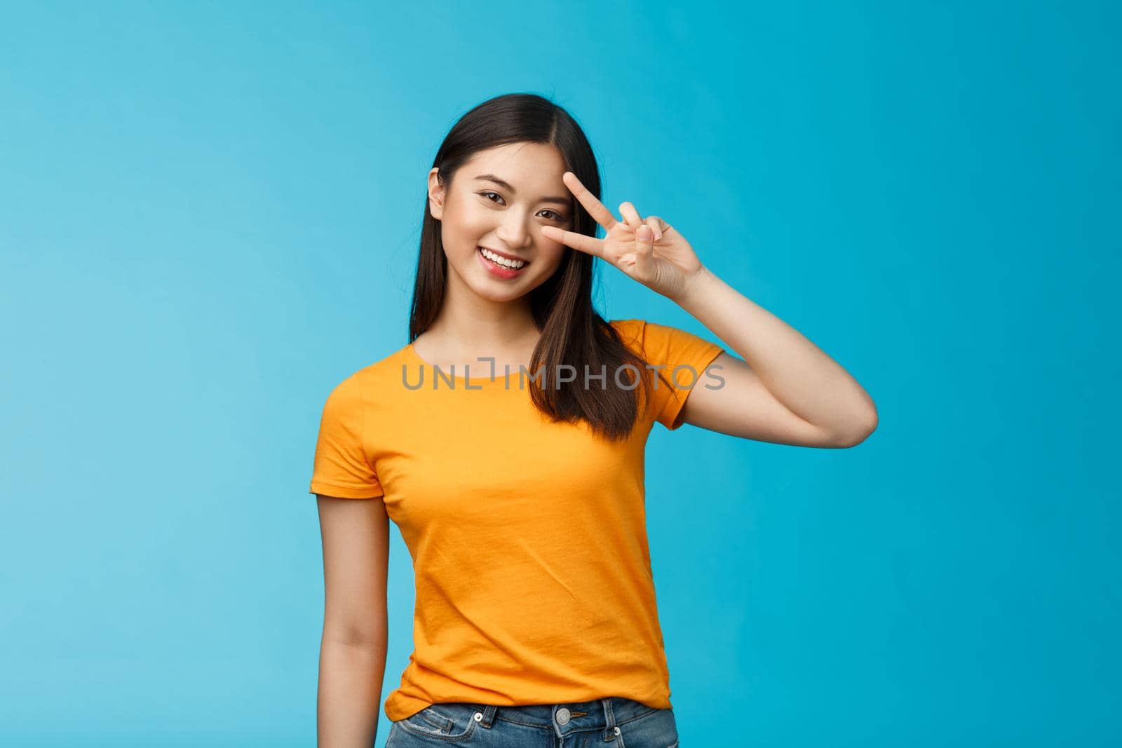 Friendly cheerful asian girlfriend look joy happiness, show peace victory sign, smiling toothy positive, enjoy lucky perfect summer sunny day, stand blue background carefree wear yellow t-shirt.