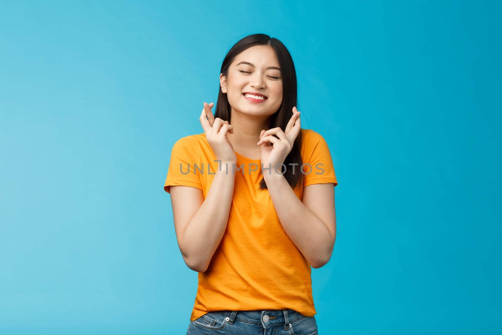 Hopeful excited asian young girl making wish cross fingers good luck, close eyes smiling believe dream come true, anticipating important event, praying good fortune, stand blue background.