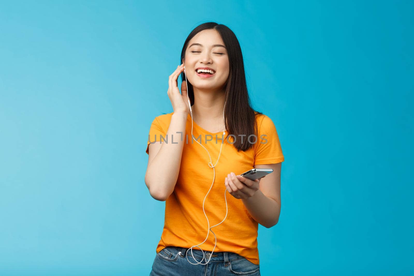 Lovely carefree cute asian girl enjoy awesome music boost mood listening favorite songs, touch earphone close eyes from pleasure delight, singing along, hold smartphone stand blue background by Benzoix