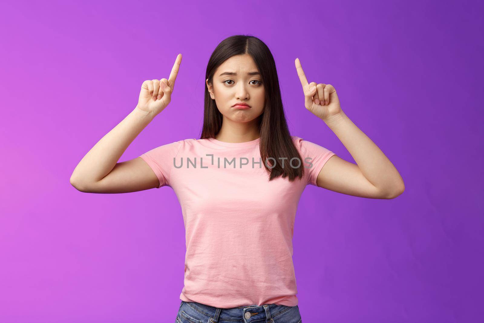 Upset moody sad asian girl look jealous, pouting distressed, pointing up indicating index fingers top advertisement regret missing chance, standing disappointed purple background displeased.