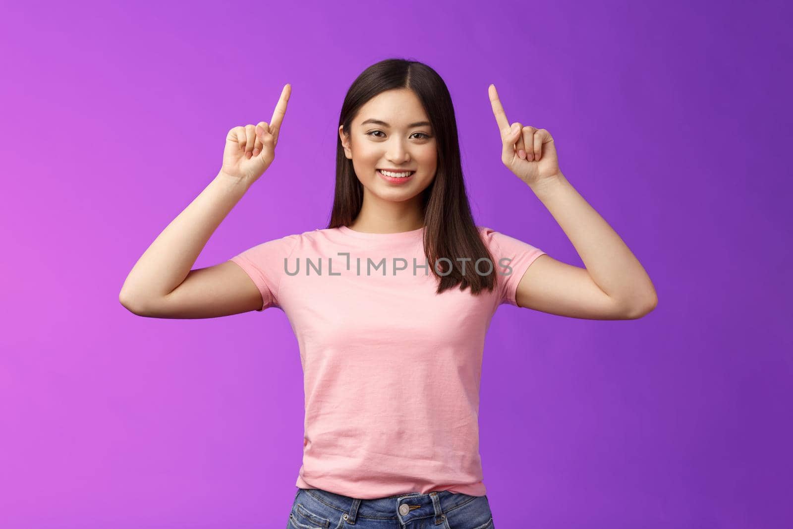 Cute assertive dark-haired asian woman wear pink t-shirt raise hands up, showing promo, pointing fingers up, grinning, inviting use link, try-out product, give direction, stand purple background.