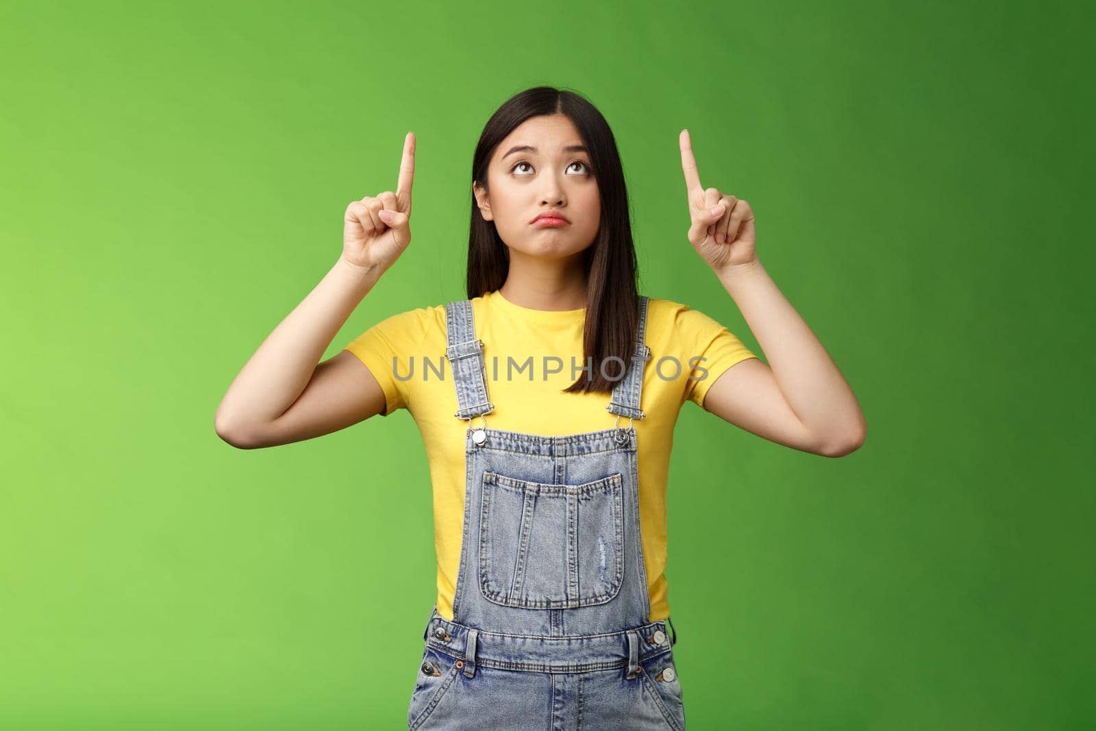 Silly upset cute asian girl look pointing up sad, gloomy watching top side, grimacing displeased, dreaming have new product, regret not have money effort cool thing, stand green background by Benzoix