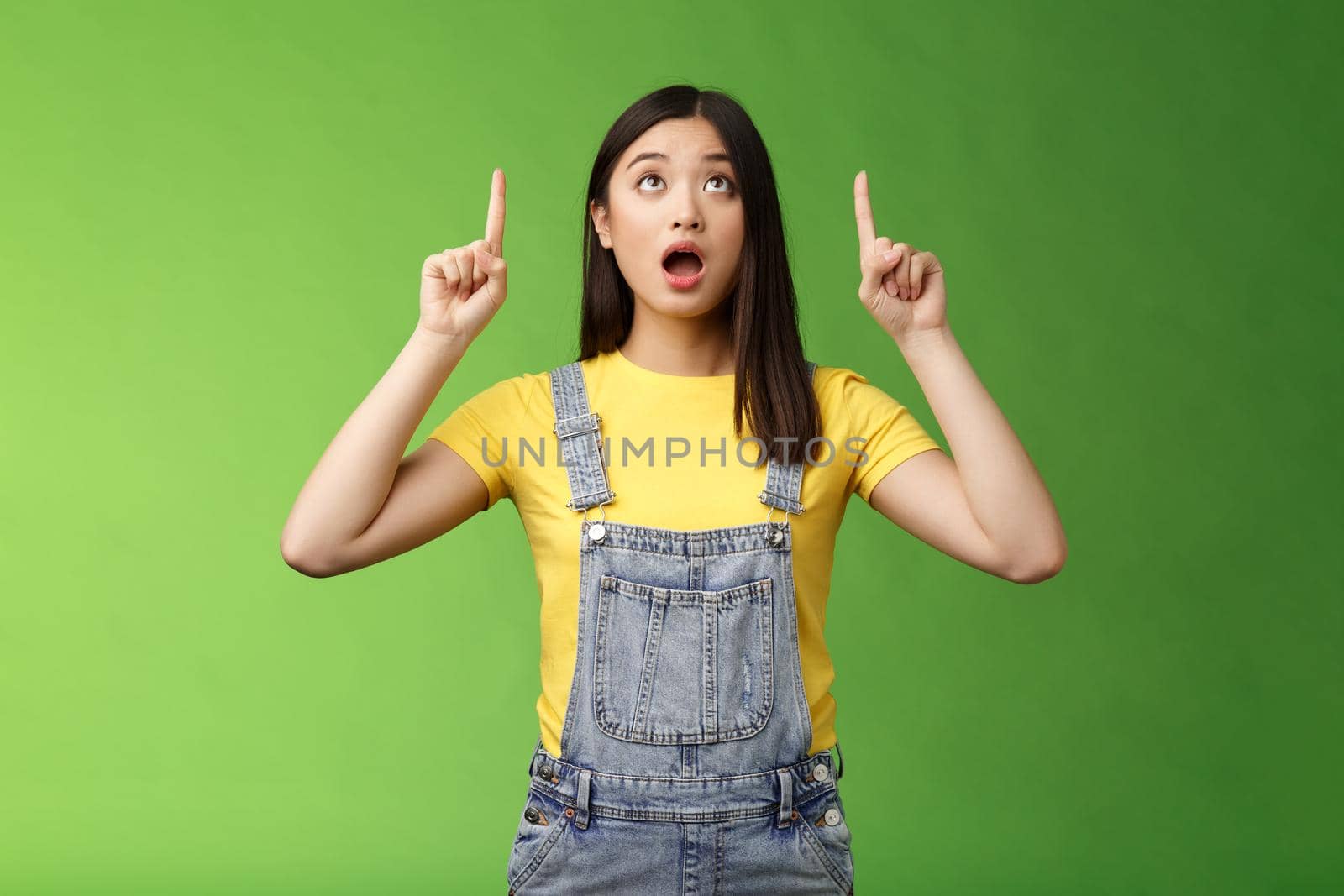 Omg look shocking price fall. Stunned speechless amazed asian girl drop jaw, wondering what happening upstairs, pointing lookin up impressed, staring surprised stand green background.