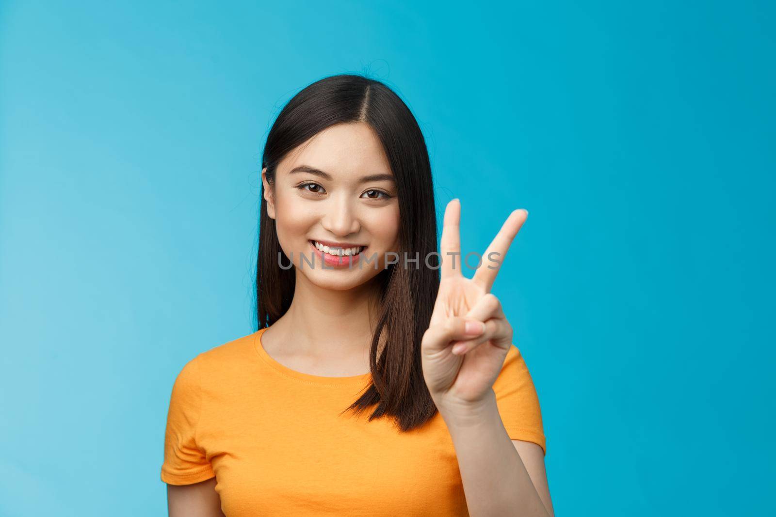 Cheerful friendly asian woman in yellow t-shirt smiling broadly, show number two, twice, win second place, smiling joyfully, make take-away order, book sits, stand blue background.