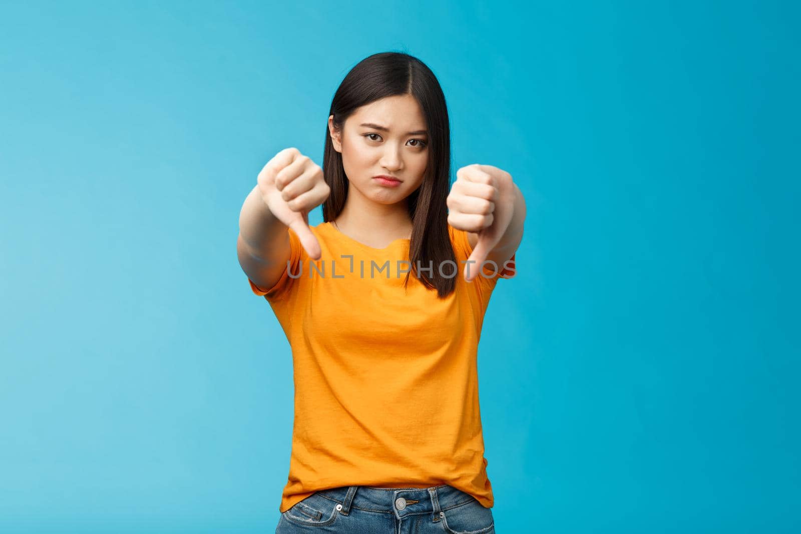 Upset gloomy asian girl with dark haircut frowning sulking sad, disappointed, show thumbs-down grimacing, give negative opinion, judging bad uninteresting movie, stand blue background.