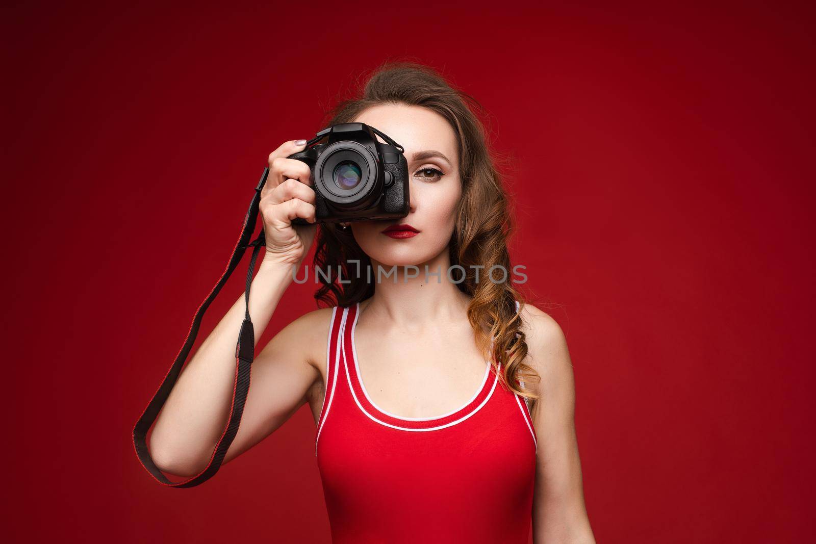 A beautiful bright woman with a camera in her hands. A woman on a red background in a red swimsuit poses with a camera. by StudioLucky