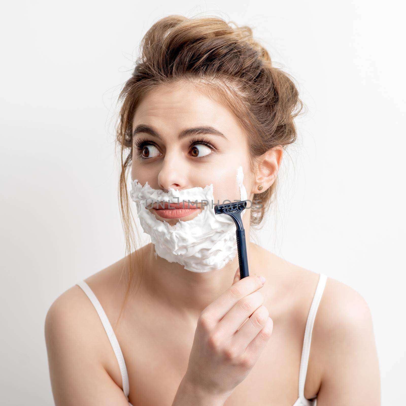 Beautiful young caucasian woman shaving her face by razor on white background. Pretty woman with shaving foam on her face