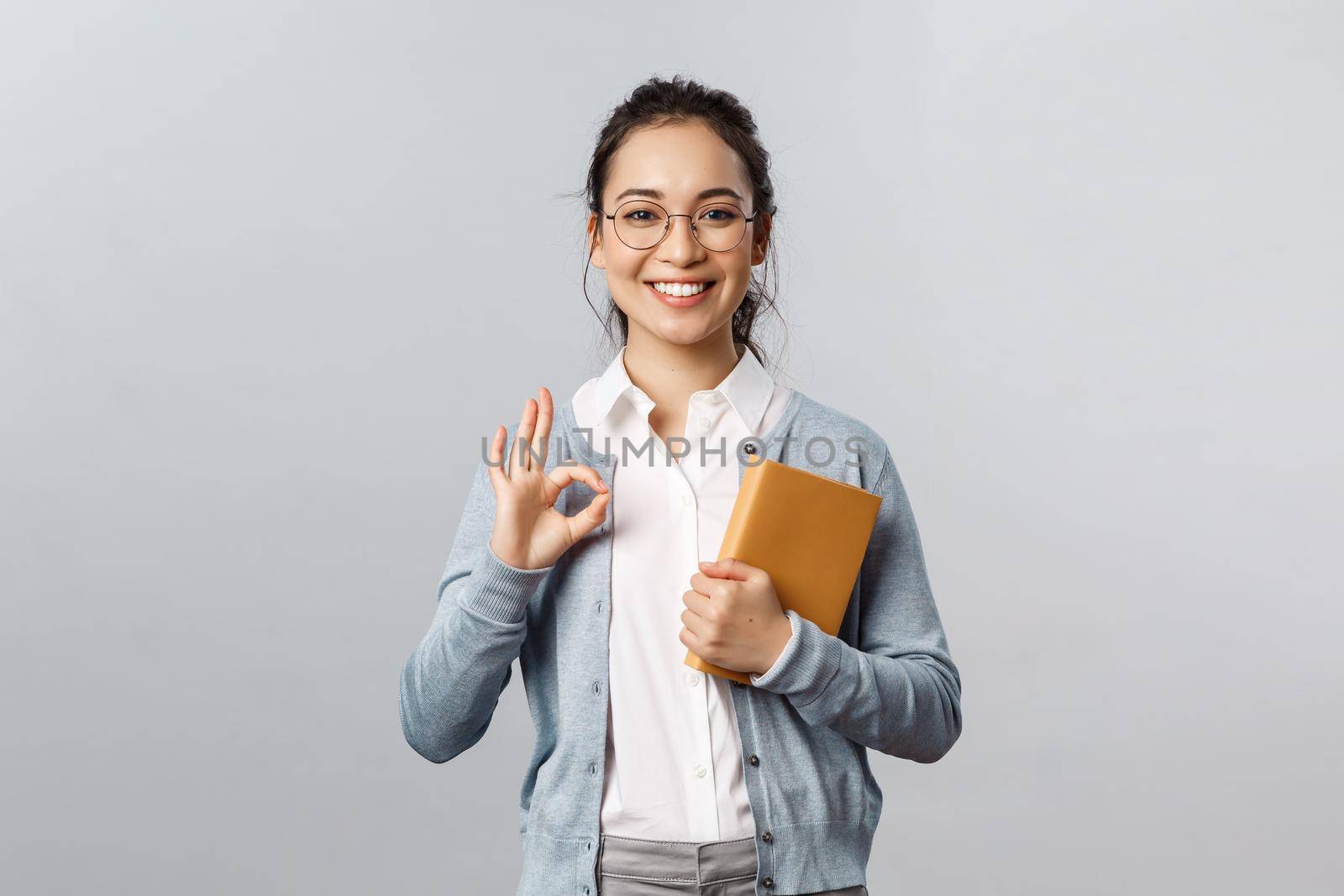 Education, teachers, university and schools concept. Portrait of young satisfied female employee or student rate good product, recommend it, show okay guarantee sign, hold planner or notebook by Benzoix
