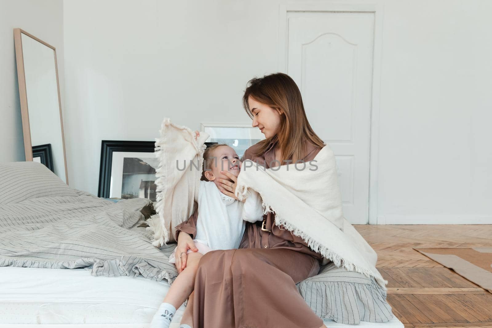 Smiling mother hugging her little daughter, sitting on a cozy bed in the bedroom. people and family concept.