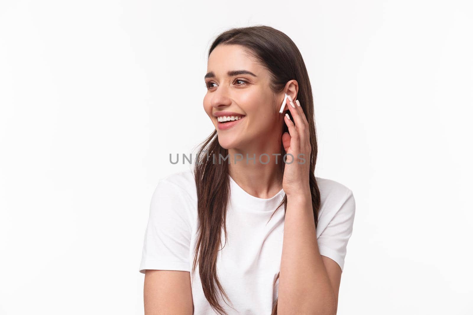 Close-up portrait of happy charming young woman with long hair, looking away smiling and laughing joyfully, changing song touching her wireless earbud, stand white background.
