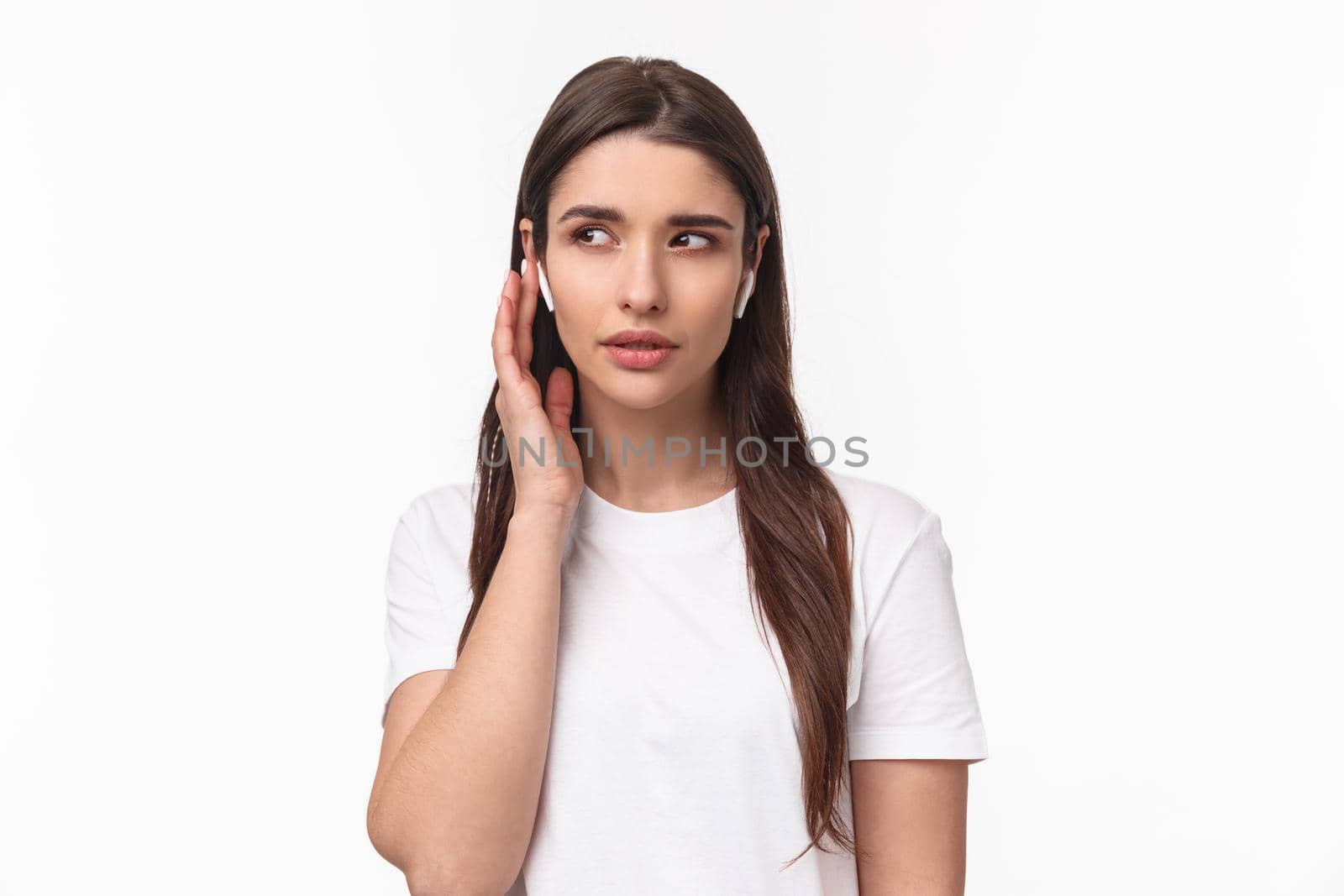 Close-up portrait of young caucasian woman in t-shirt, wearing wireless headphones, listen closely, fixing volume, touching earphone to answer call, standing white background enjoying music.