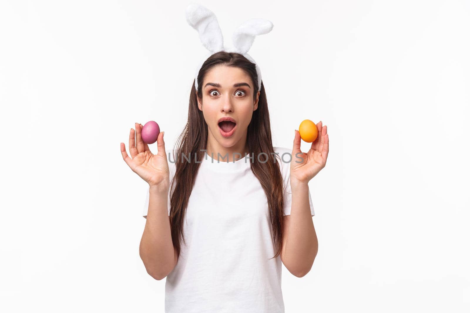 Portrait of surprised and amazed young brunette girl holding two colored eggs, wearing rabbit ears, party outfit as celebrating Easter day, enjoying holy holiday, white background by Benzoix