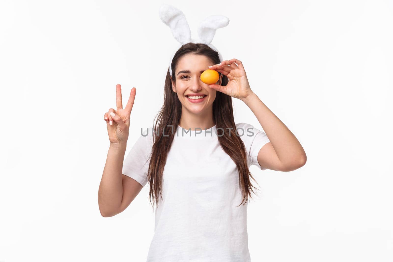 Portrait of peaceful and cheerful young cute female in white t-shirt, showint peace sign and hold colored egg over eye, smiling happy, enjoying traditional holiday celebration, Easter day.