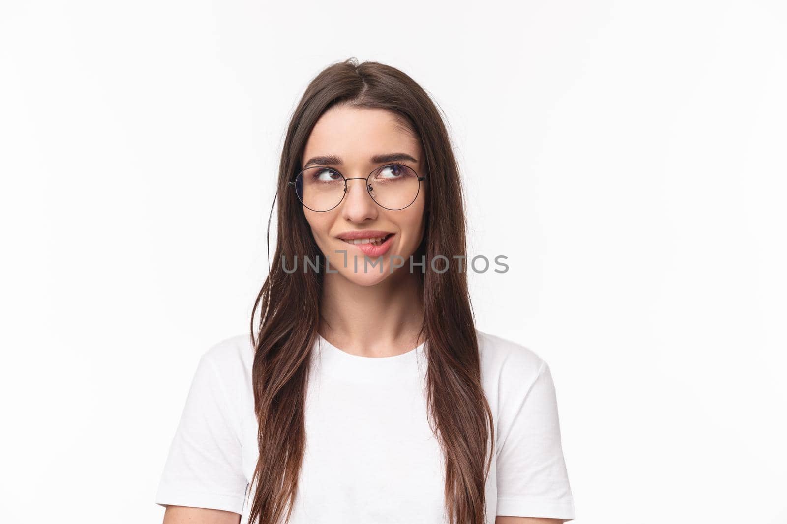 Close-up portrait of sassy and cute young brunette girl, biting lip tempting do something, having plan, look up thoughtful, standing white background, imaging or daydreaming. Copy space