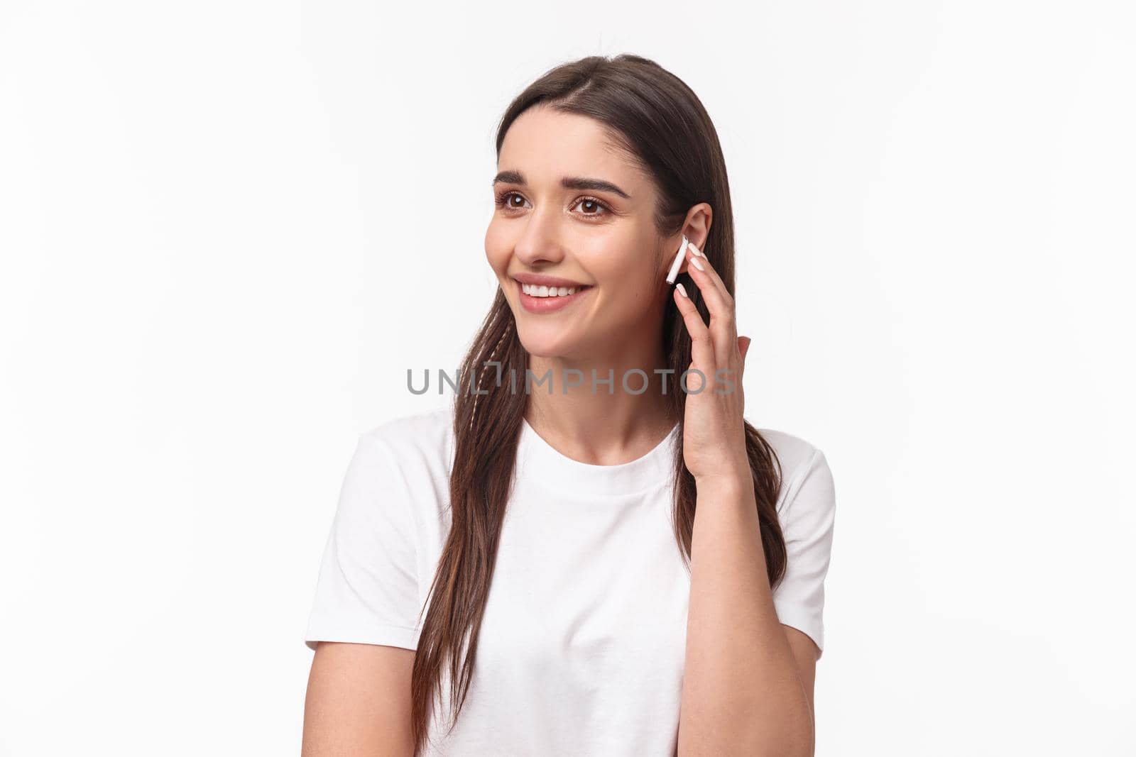 Close-up portrait of attractive smiling young brunette woman talking on phone, using wireless earphone to contact friend, looking away carefree, listening music in headphones, white background.