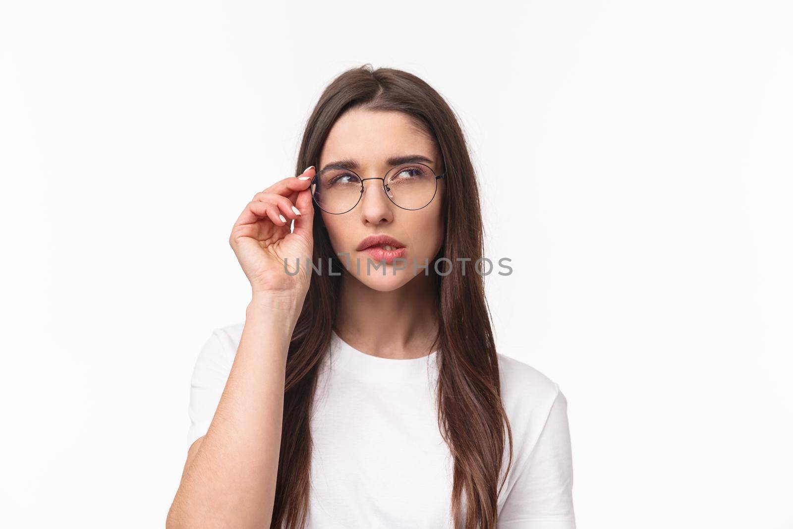 Close-up portrait of smart and thoughtful beautiful young girl in glasses, thinking, squinting suspicious and biting lip look up while pondering something, have interesting idea, white background.