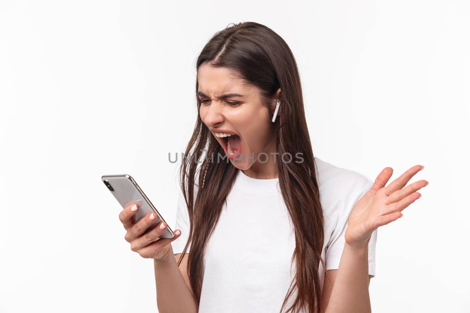 Close-up portrait of pissed-off aggressive and angry young brunette woman yelling at person while having phone call in wireless earphones, screaming at smartphone outraged.