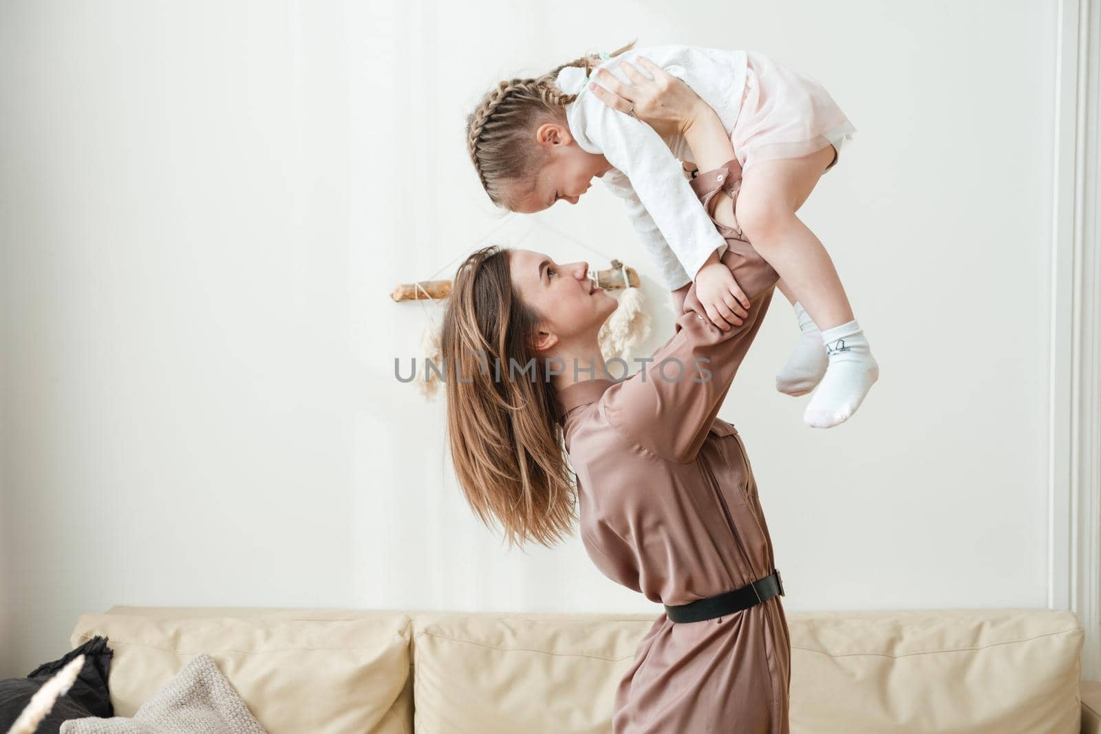 Smiling mother lifts happy little daughter into the air in the living room and plays with her