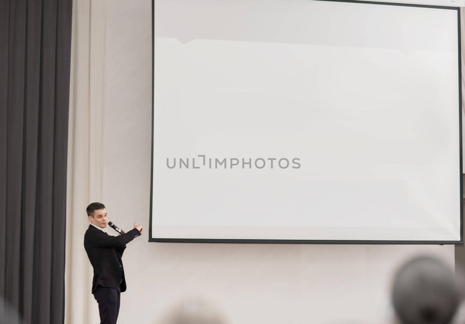 speaker conducts the business of the conference standing in front of a large white screen on the stage in the conference room by SmartPhotoLab