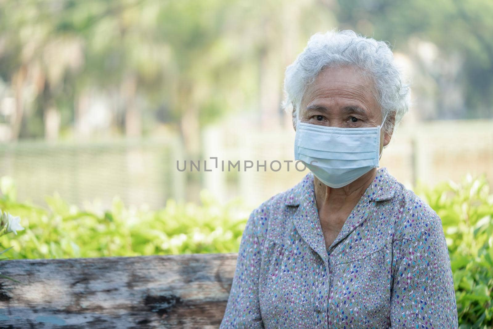 Asian elderly woman sitting bench and wearing face mask for protect safety infection Covid-19 Coronavirus social distancing in park.