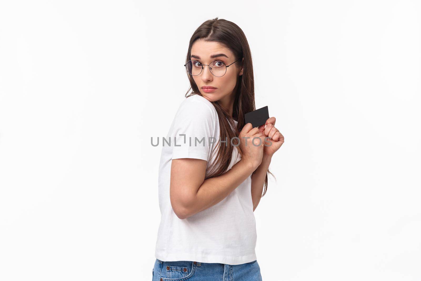 Portrait of greedy and funny young woman unwilling to give her credit card, hiding it and looking with disbelief and reluctance, have money but dont like share, standing white background.