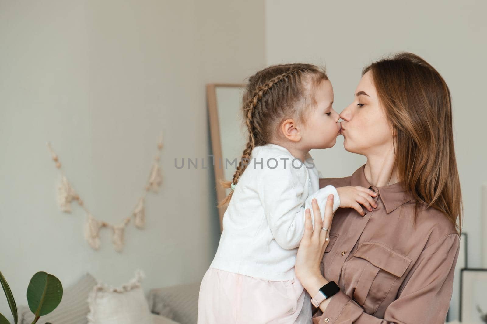 A little girl kisses her mother at home in a modern bright bedroom. The concept of parenthood, love and care. An adult woman holds her daughter in her arms and hugs her. Mothers Day.