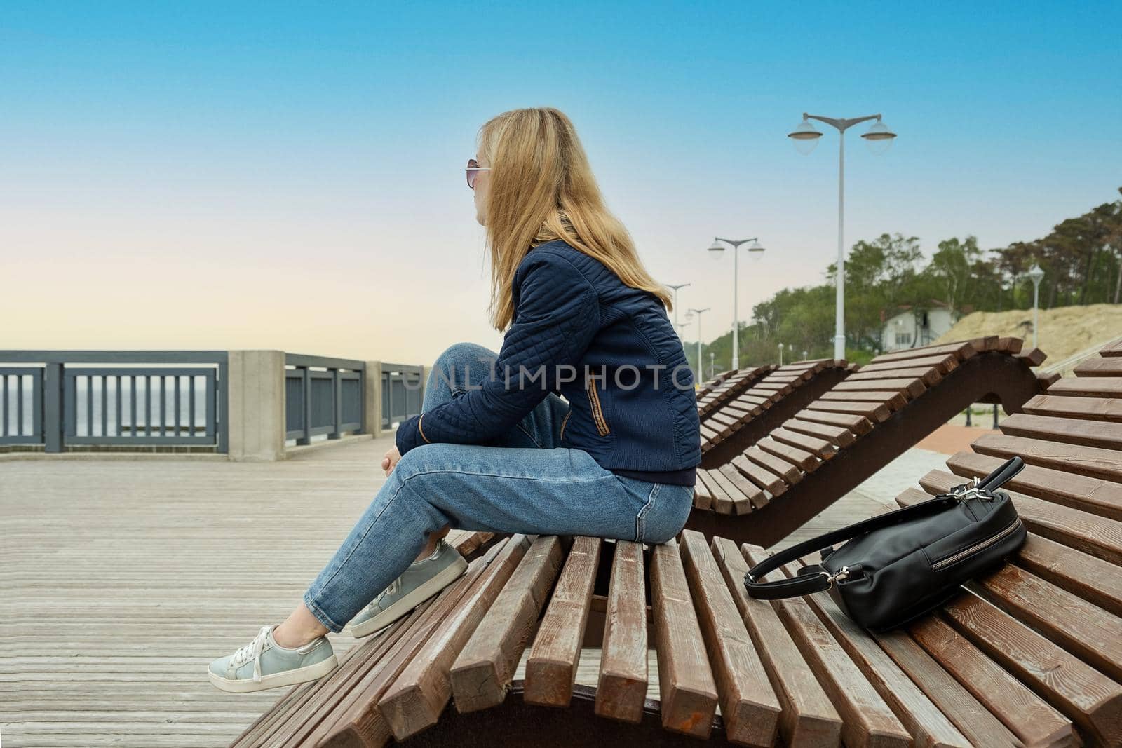 A young lonely blonde woman on an empty city embankment rests sitting on a sun lounger on a spring or autumn evening and looks into the distance. The girl misses the warmth and summer.