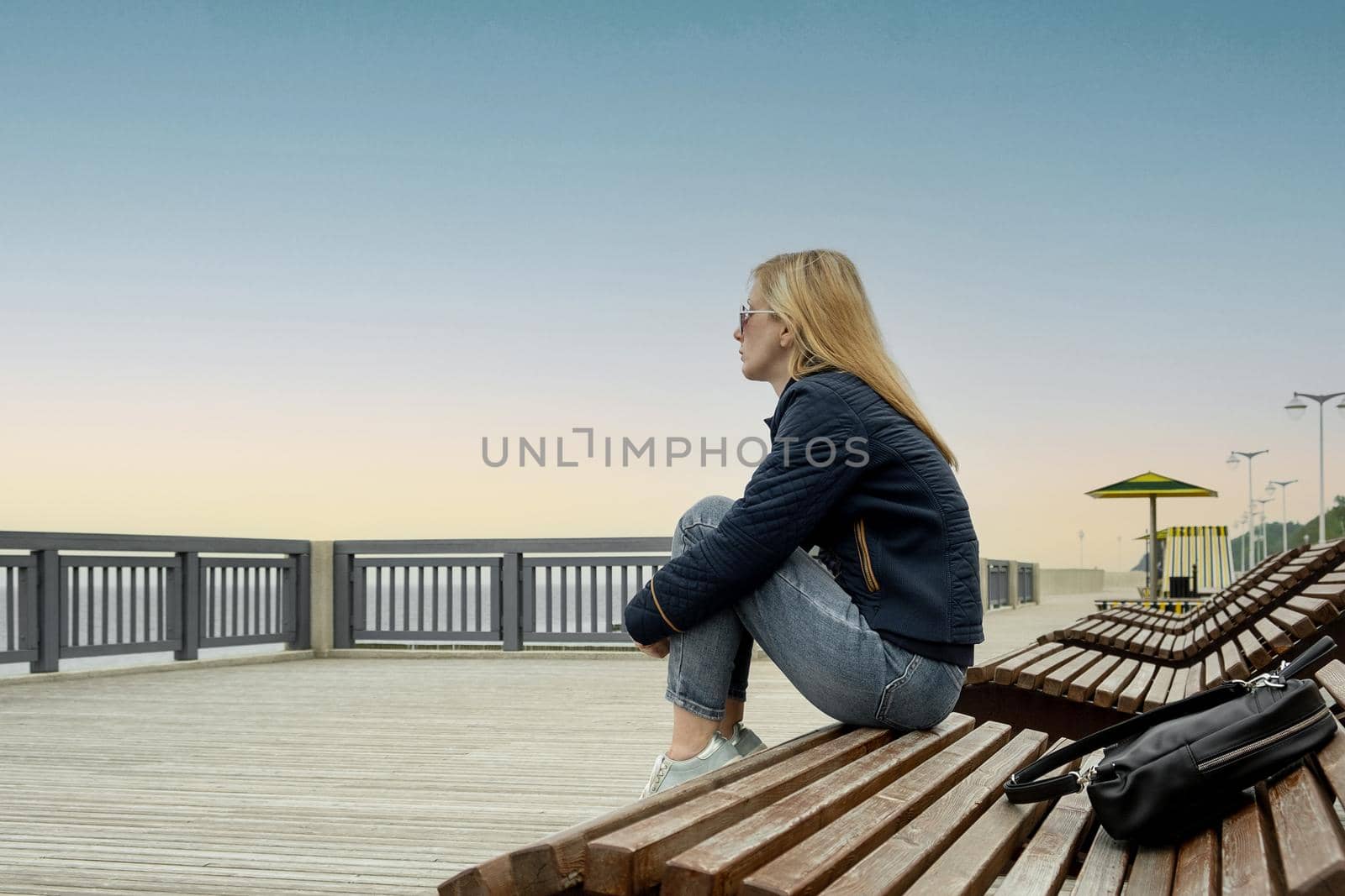 A young lonely blonde woman on an empty city embankment rests sitting on a sun lounger on a spring or autumn evening and looks into the distance. The girl misses the warmth and summer.
