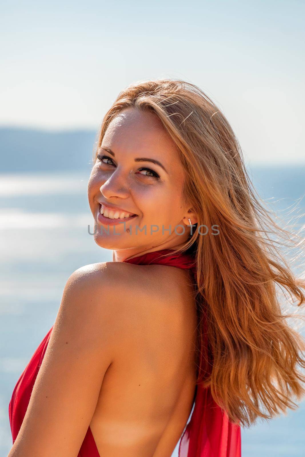 Smiling young woman in a red dress looks at the camera. A beautiful tanned girl enjoys her summer holidays at the sea. Portrait of a stylish carefree woman laughing at the ocean