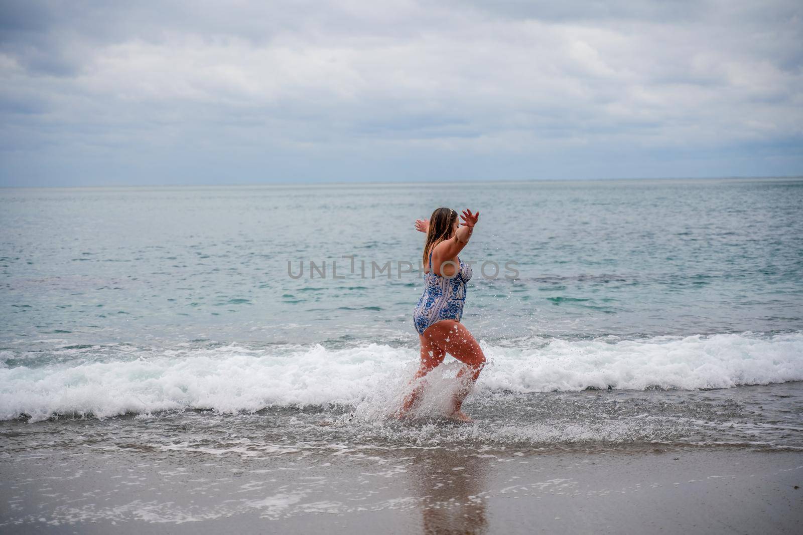 A plump woman in a bathing suit enters the water during the surf. Alone on the beach, Gray sky in the clouds, swimming in winter