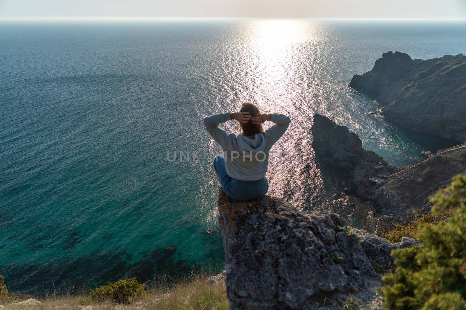 Woman tourist enjoying the sunset over the sea mountain landscape. Sits outdoors on a rock above the sea. She is wearing jeans and a blue hoodie. Healthy lifestyle, harmony and meditation by Matiunina