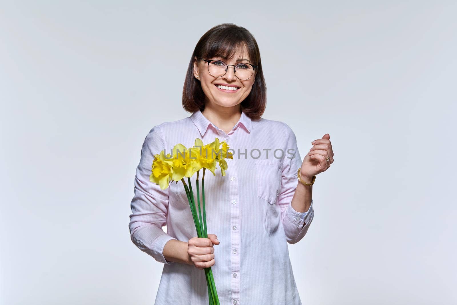 Portrait of beautiful middle aged woman with bouquet of flowers looking at camera on light studio background. Charming smiling mature woman. Beauty, holiday, joy, happiness, people concept