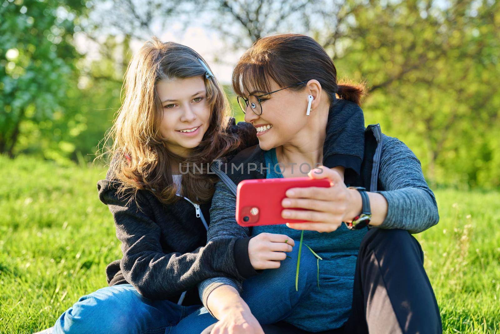 Mom and daughter 11, 12 years old having fun looking together at smartphone screen, outdoor, sitting embracing on grass. Family, leisure, lifestyle, relationship love, mother's day, motherhood concept