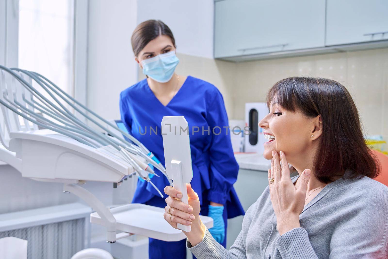 Dentist's office, woman patient looking at her teeth in the mirror. Doctor dentist near the dental chair with tools. Treatment, dental care, prosthetics, orthodontics, dentistry concept