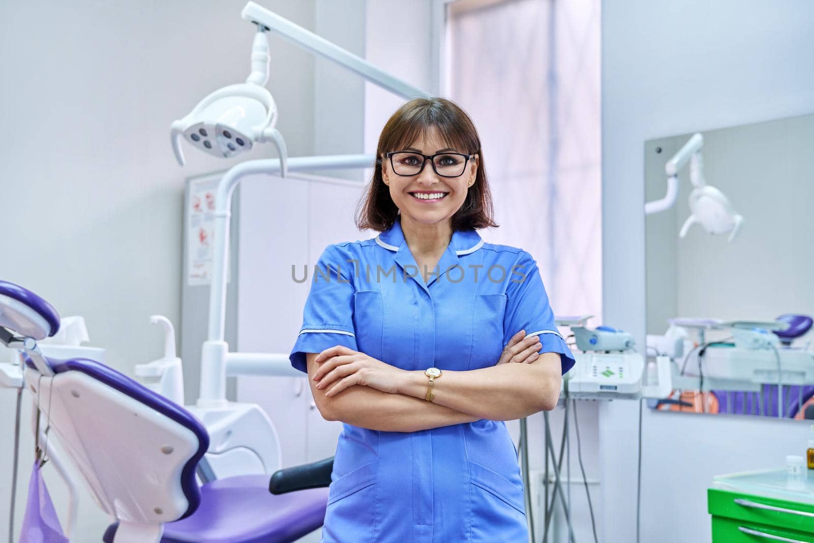 Portrait of smiling nurse looking at camera in dentistry. Confident female with folded hands, dental clinic equipment background. Dentistry, medicine, health care, profession, stomatology concept