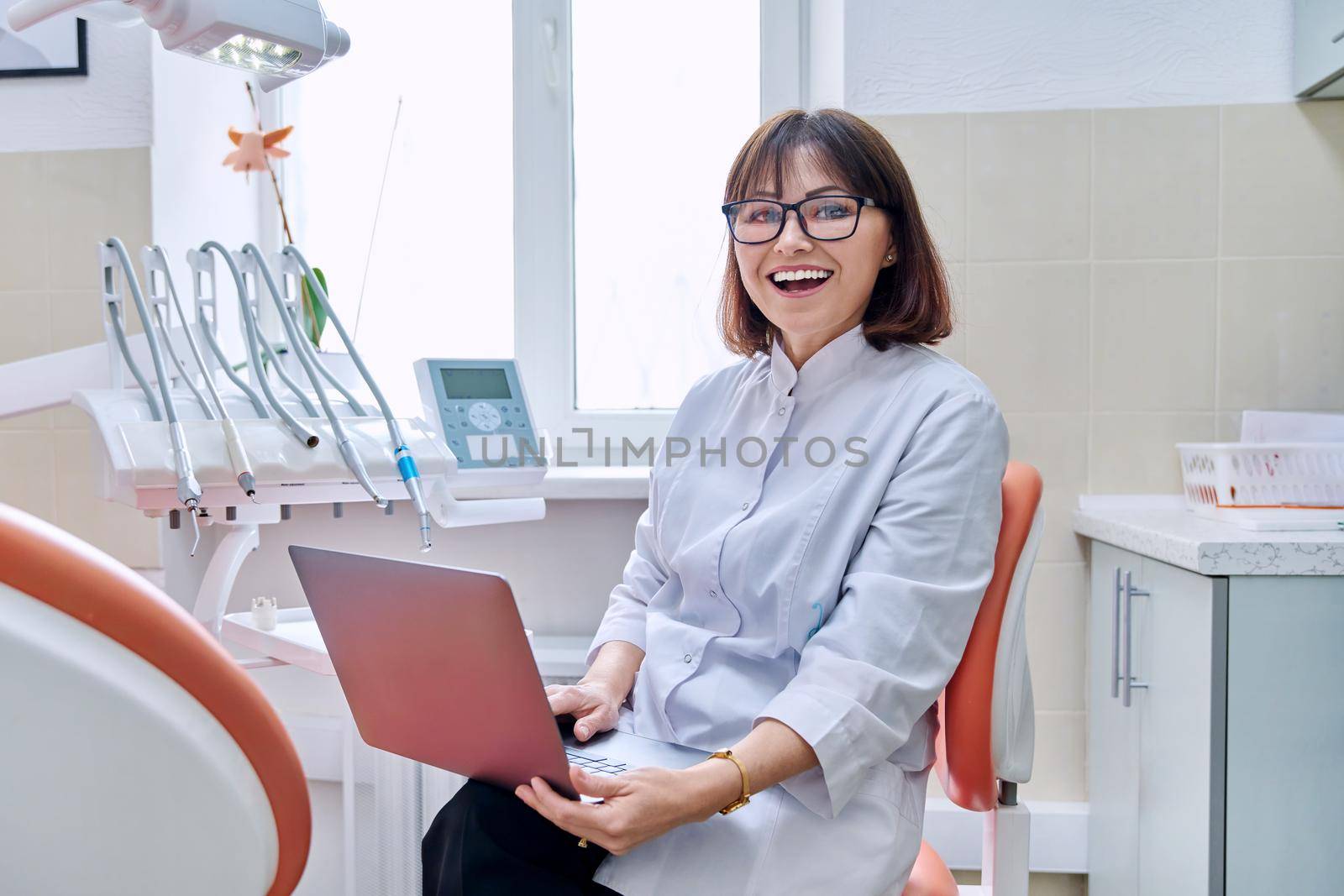 Portrait of doctor sitting in office with laptop in her hands looking at camera by VH-studio