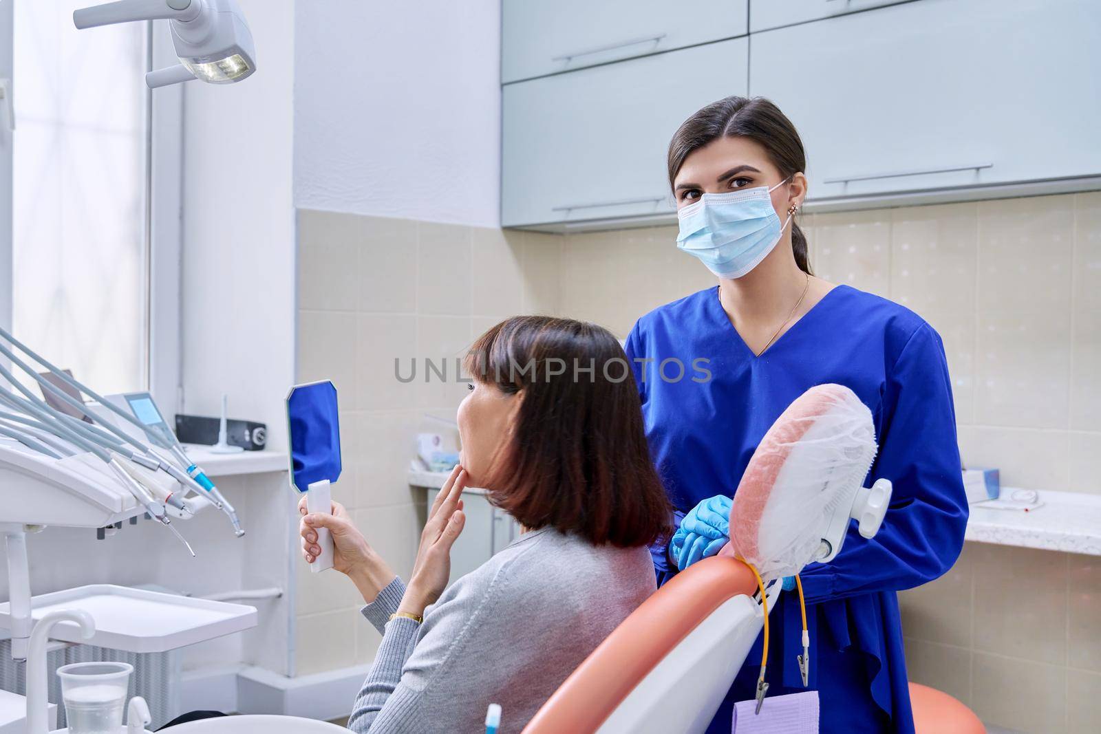 Dentist's office, woman patient looking at her teeth in the mirror. Doctor dentist near the dental chair with tools. Treatment, dental care, prosthetics, orthodontics, dentistry concept