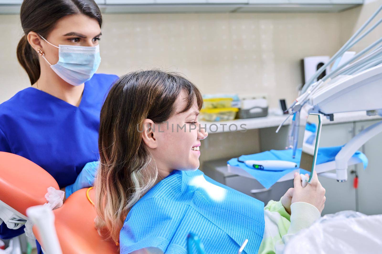 Teenage female looking at healthy teeth in mirror, in dental office, visit to dentist orthodontist. Treatment and care of teeth, teenagers, orthodontics, dentistry concept