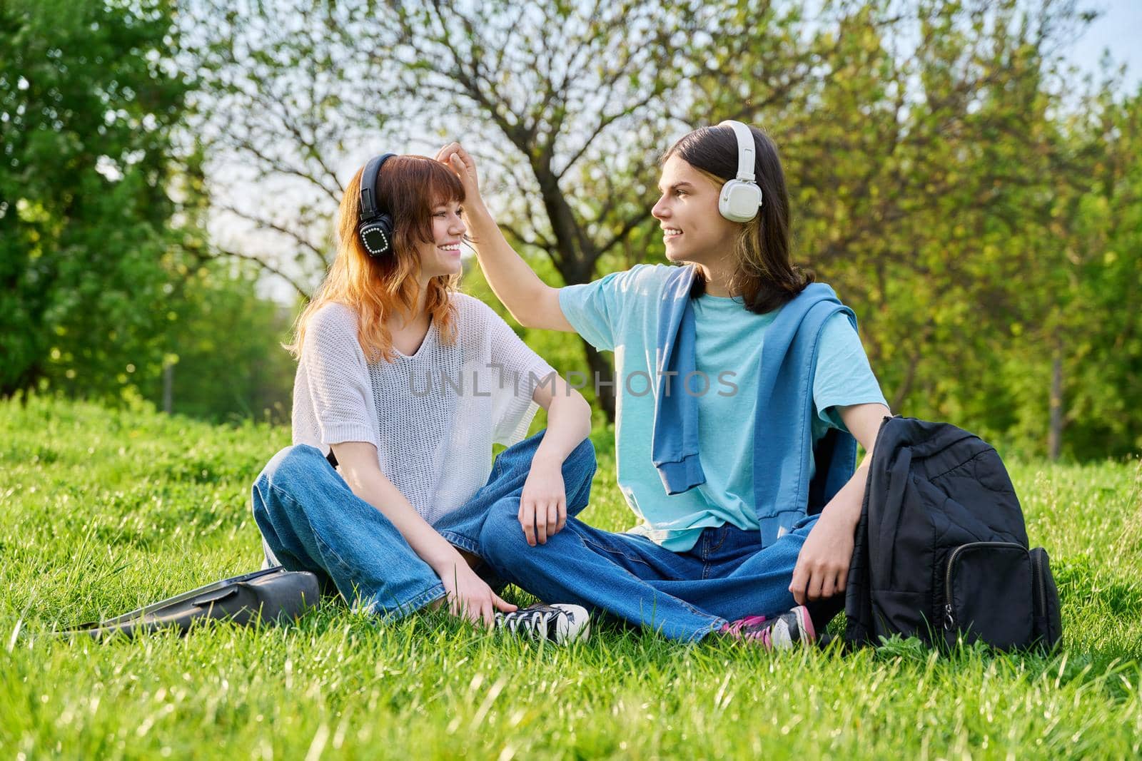 Couple of friends guy and girl 17, 18 years old sitting on grass. Students in headphones with backpacks sitting on campus lawn, talking and laughing. Teens, youth, friendship, education, young people