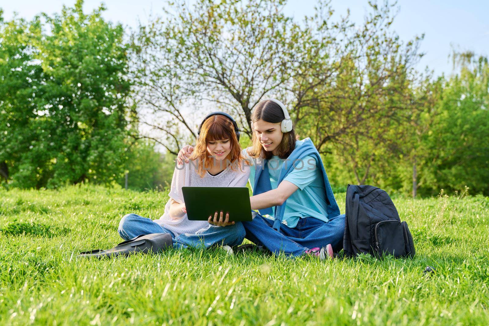 Two friends students, guy and girl in headphones with backpacks, looking at laptop, sitting on grass. High school, college, communication, friendship, education, youth concept