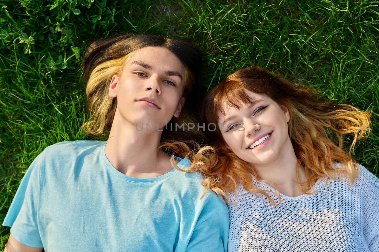 Couple of teenagers friends looking at camera on green grass background. Happy guy and girl 17, 18 years old lying on lawn, top view. Youth, friendship, lifestyle, young people concept