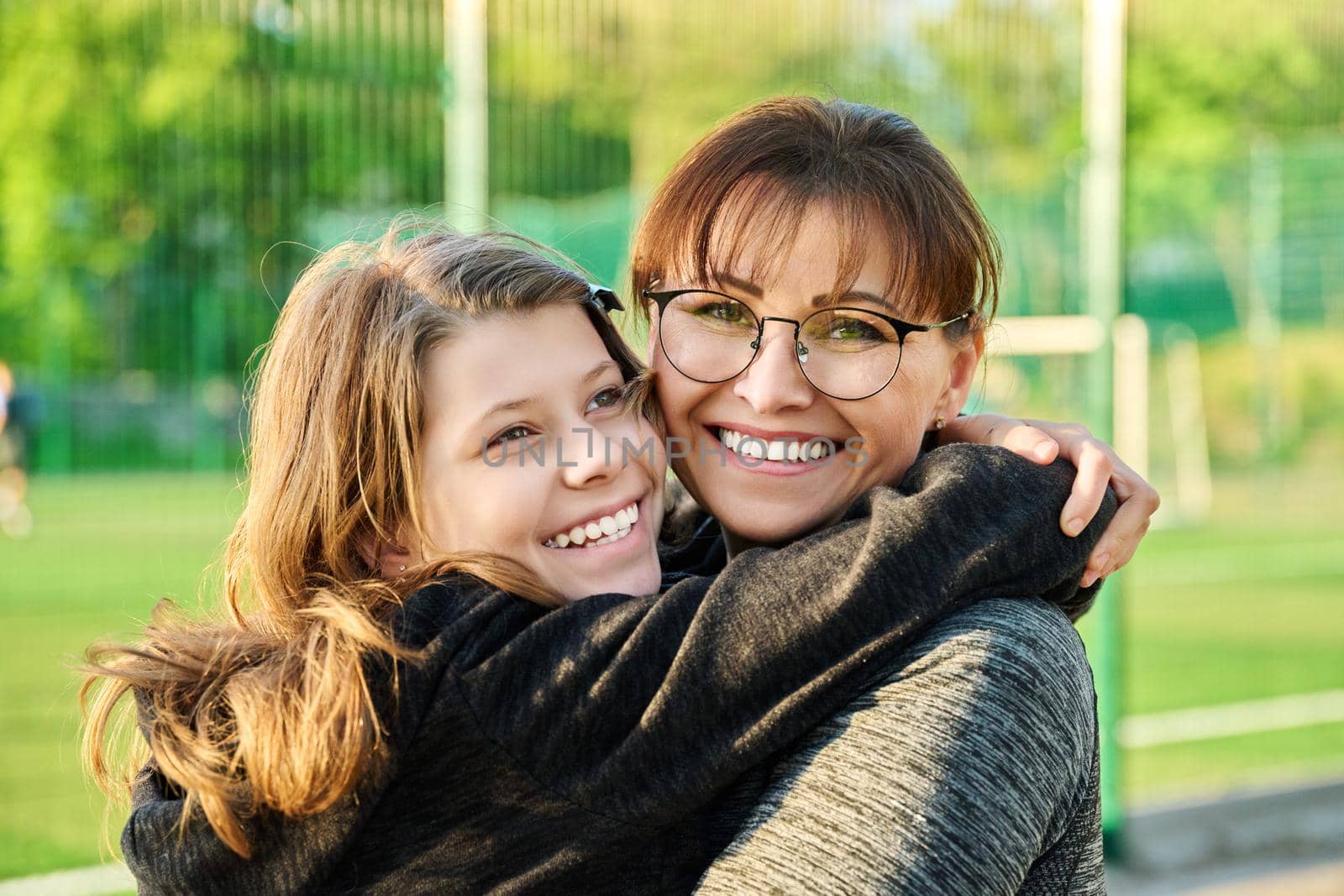 Portrait of happy mom and preteen daughter hugging together outdoor, near sports stadium, football field. Family, happiness, leisure, lifestyle, relationship, love, mother's day, motherhood concept