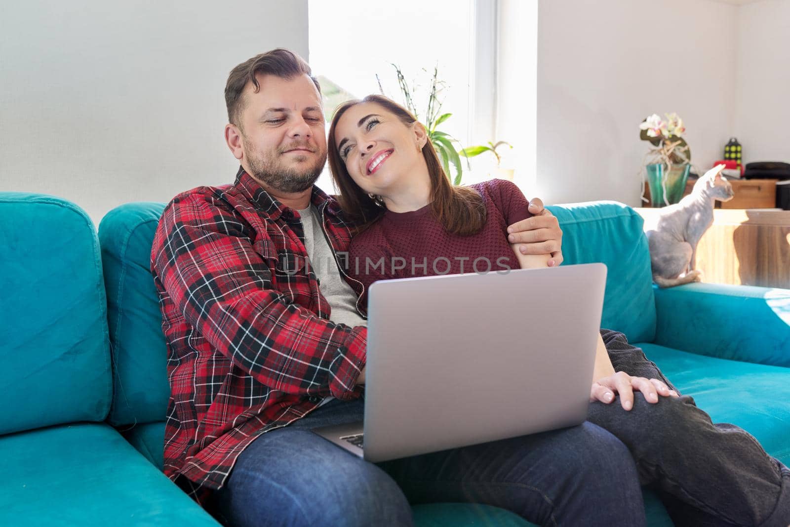 Happy smiling middle aged family couple looking at laptop screen together while sitting on sofa at home with pet cat. Relationships, lifestyle, family, leisure, technology, rest, people 40s concept