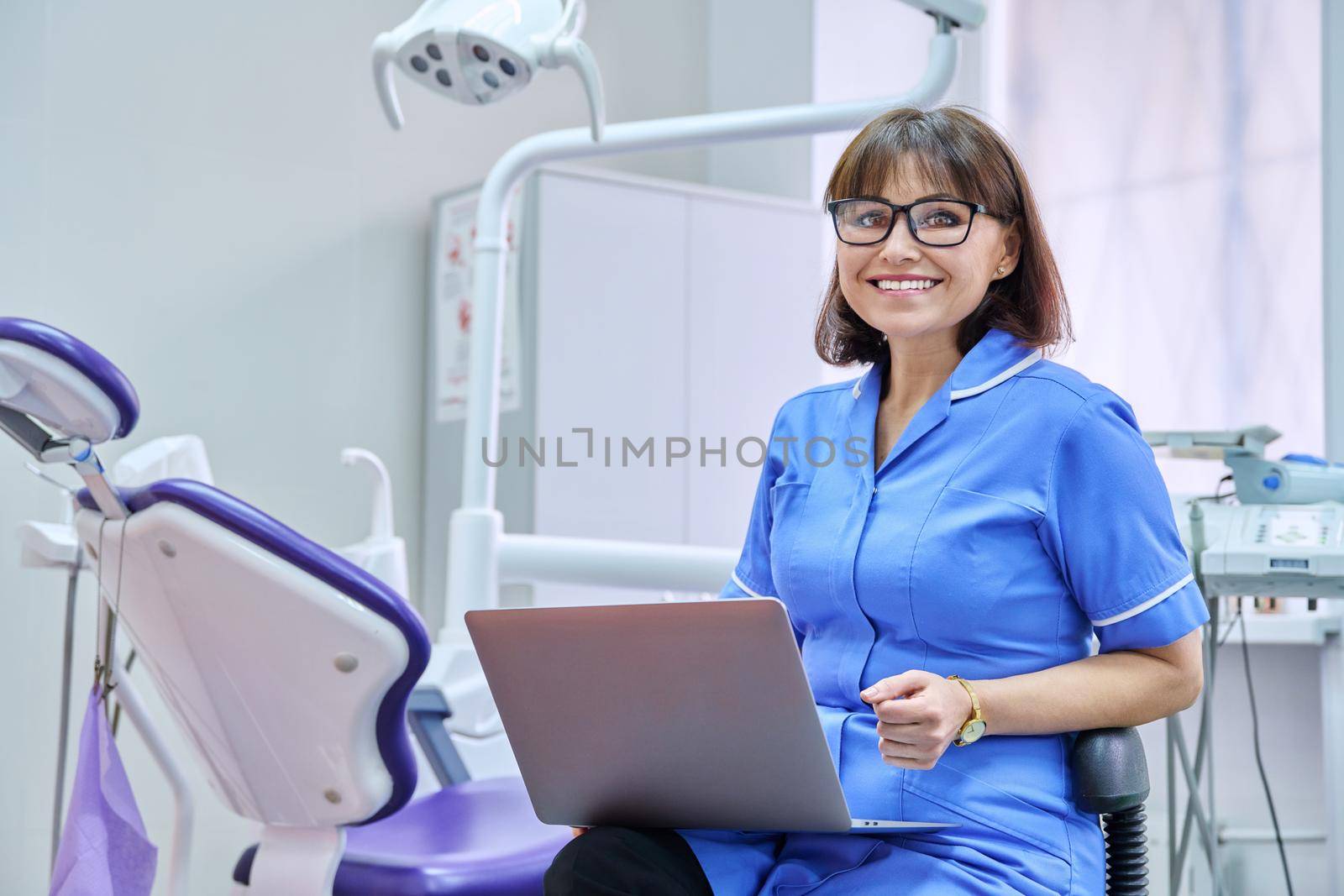 Portrait of dentist nurse sitting in office with laptop in her hands looking at camera by VH-studio