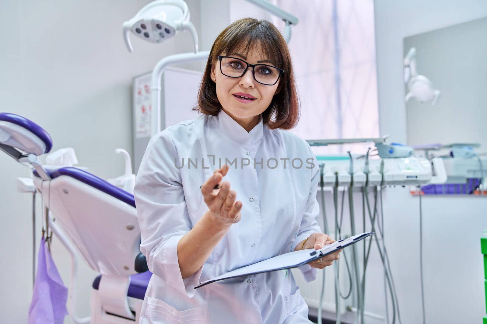 Talking consulting female doctor dentist looking at webcam, making video call chat conference, notes on clipboard, in dental office. Online technology, telemedicine, help medicine, health care concept