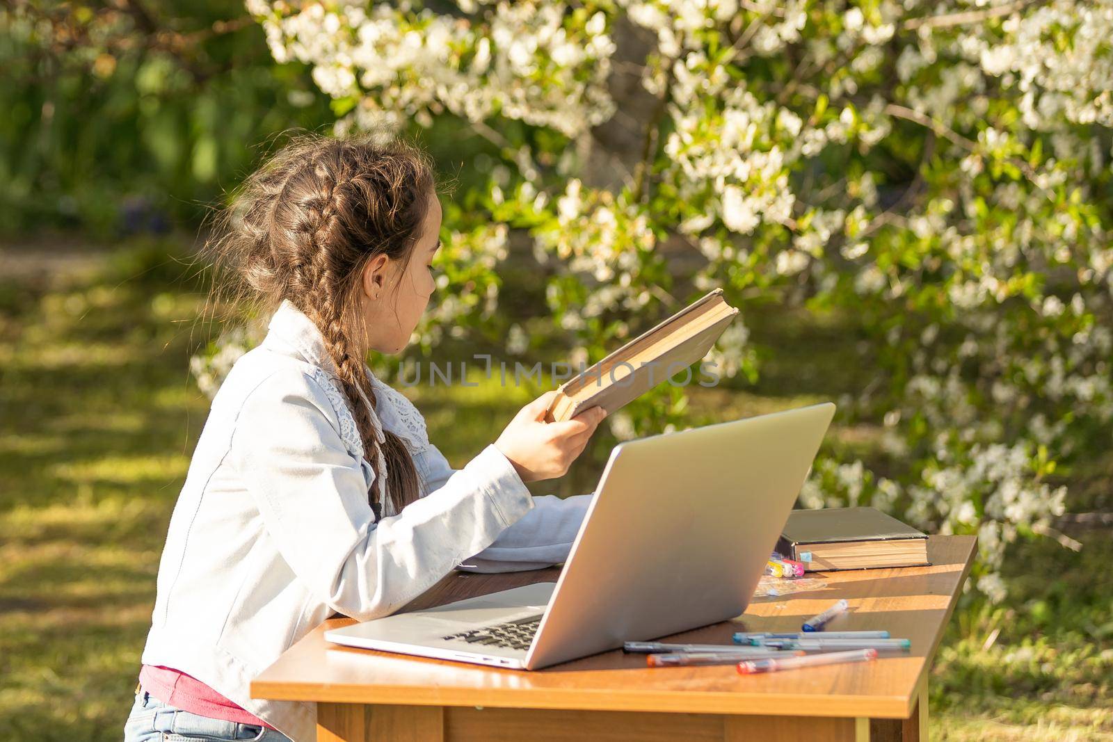 schoolgirl sits outdoors, types on keyboard of laptop. little caucasian girl sitting at table at veranda of cafe, studying with computer, doing homework. distant learning, online education by Andelov13