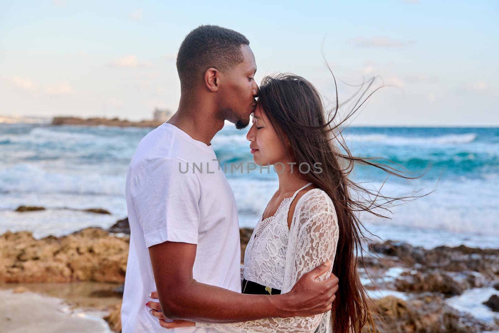 Kiss of loving young couple, sea sky background. Beautiful multicultural couple, profile view, honeymoon at seaside resort, vacation together. Love, relationship, tourism travel, nature beauty people