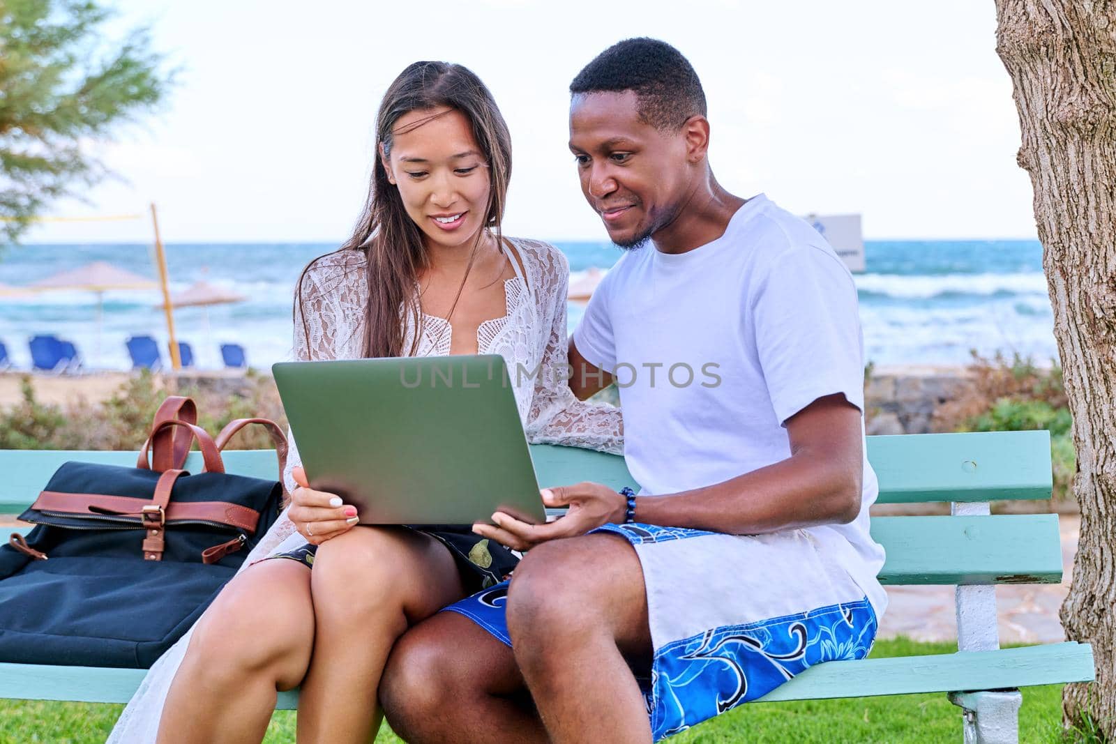 Young couple looking at laptop screen, sitting on bench on the beach, sea nature background. Lifestyle, vacation, summer, family, leisure, relationship, people concept