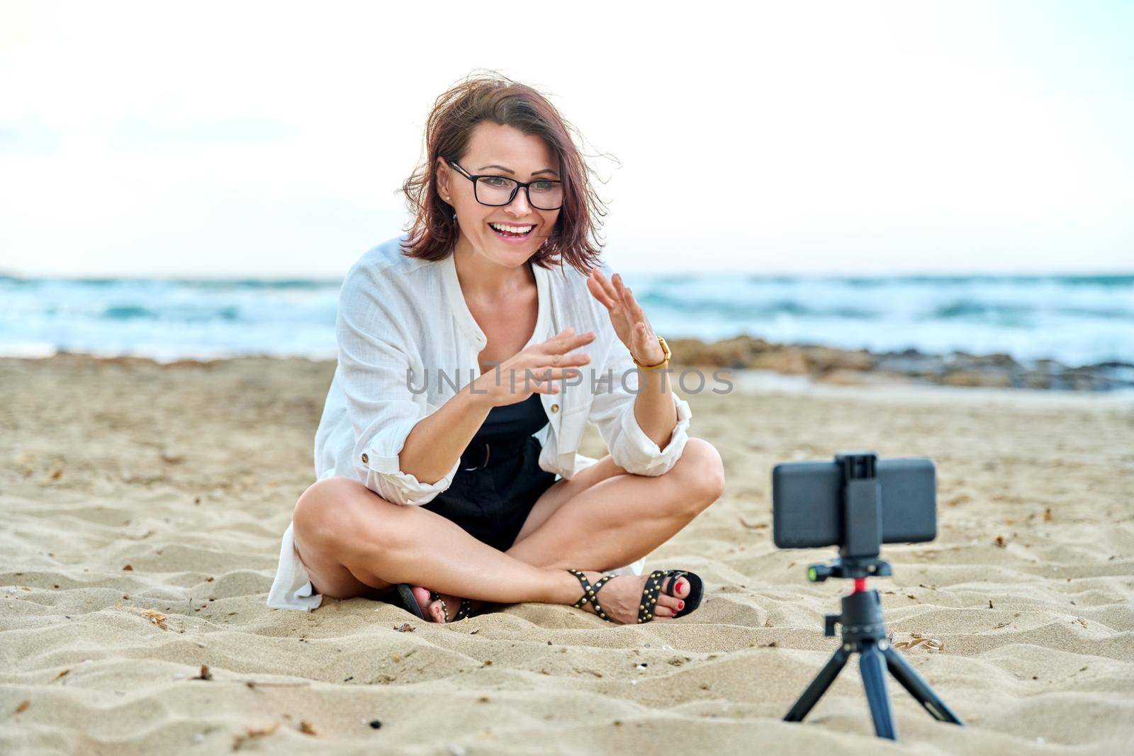 Middle aged woman sitting on beach with smartphone using video call by VH-studio